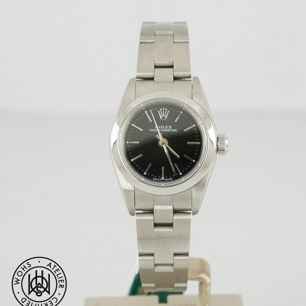 Rolex Oyster Perpetual stainless steel watch with a black dial on a display stand, plus a circular certification seal in the bottom left corner.