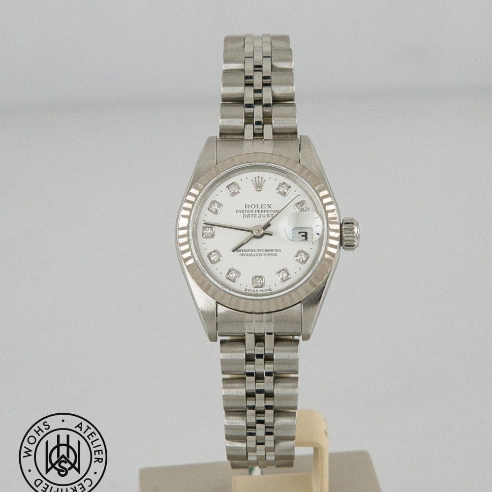 Silver Rolex Datejust wristwatch with diamond hour markers on white dial and a brushed metal bracelet, date at 3 o'clock magnified by cyclops lens, mounted on display stand.