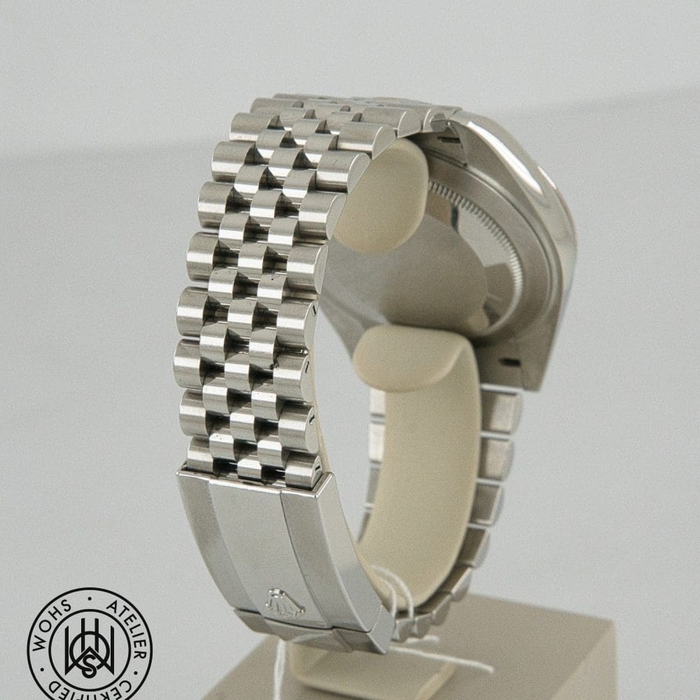 Close-up of a stainless steel watch bracelet on a display stand, showing the links and clasp with a circular certification mark in the bottom-left corner. DSC03103