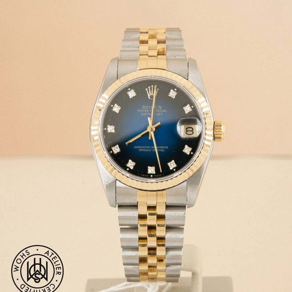 Two-tone Rolex Datejust wristwatch with a blue dial, diamond hour markers, and a date window at 3 o'clock on a display stand.