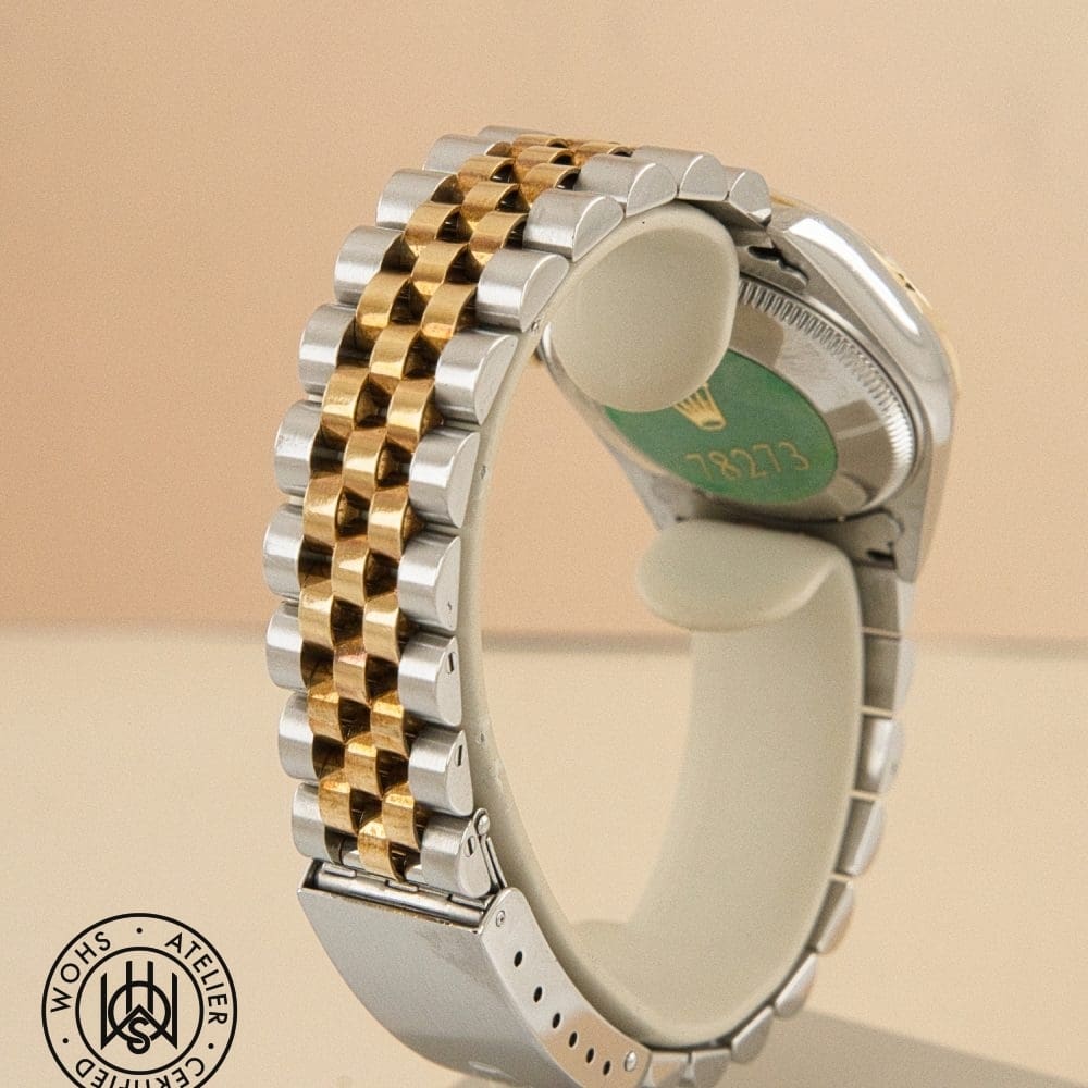 Close-up of a two-tone gold and silver metal watch band curved into a loop, with the watch back visible on a peach background. DSC03155