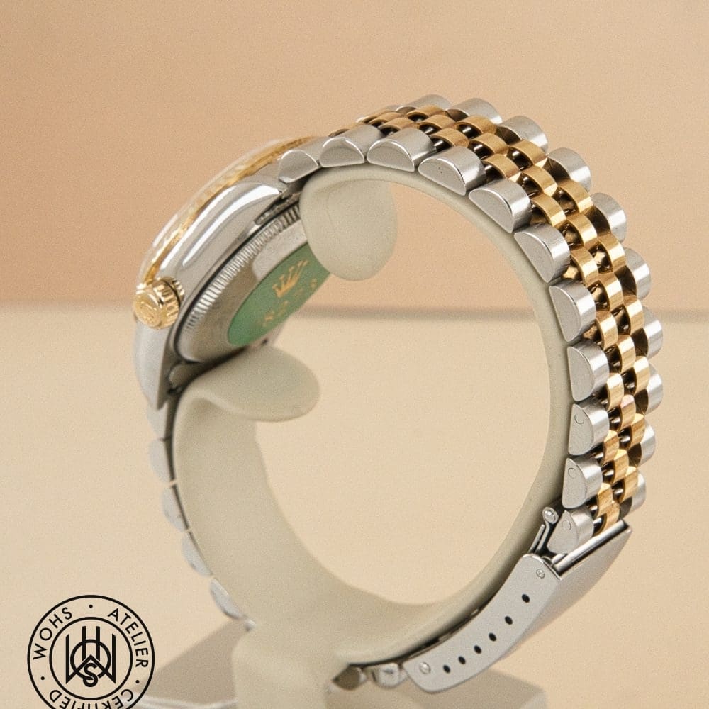 Close-up of a two-tone metal watch bracelet (silver and gold links) curved around a display stand with a circular watermark in the bottom-left corner. DSC03156