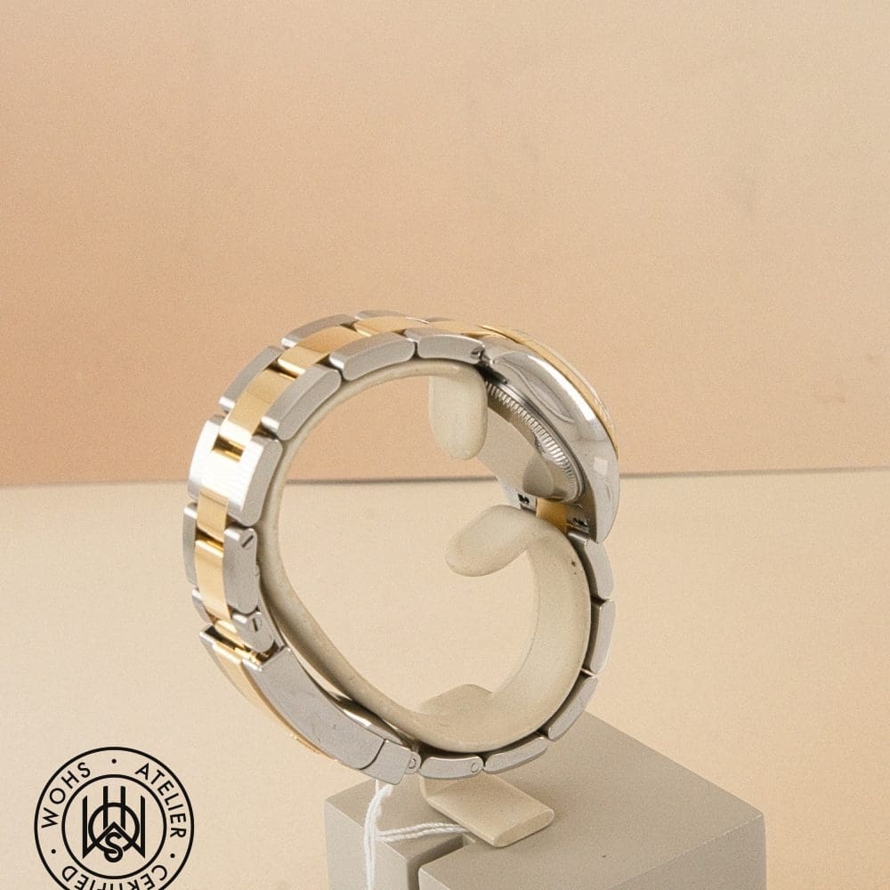 Two-tone metal watch bracelet on a display stand against a beige background, with a circular logo in the bottom-left corner. DSC03249