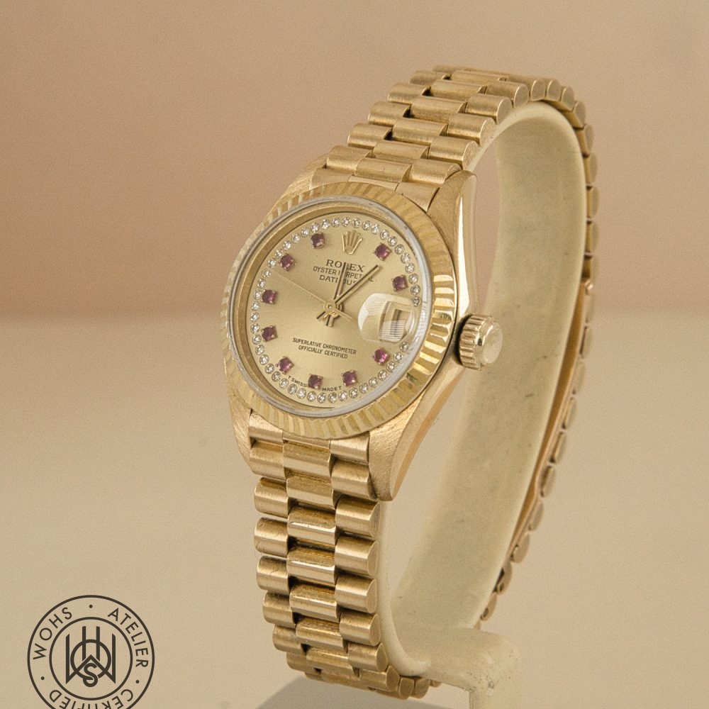 Gold Rolex Datejust wristwatch with diamond markers on a beige background, displayed on a stand; watermark bottom-left. Rolex Ladies Datejust reference 69178 - Diamond and Ruby Dial Pave 18ct Gold Dial 1