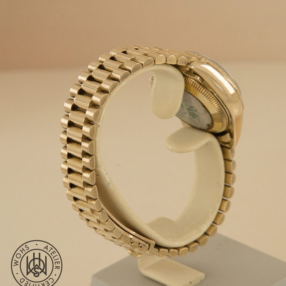Gold-toned wristwatch with a segmented bracelet displayed on a stand, curved to reveal the back case and a circular seal in the bottom-left corner. Rolex Ladies Datejust reference 69178 - Diamond and Ruby Dial Pave 18ct Gold Dial 4
