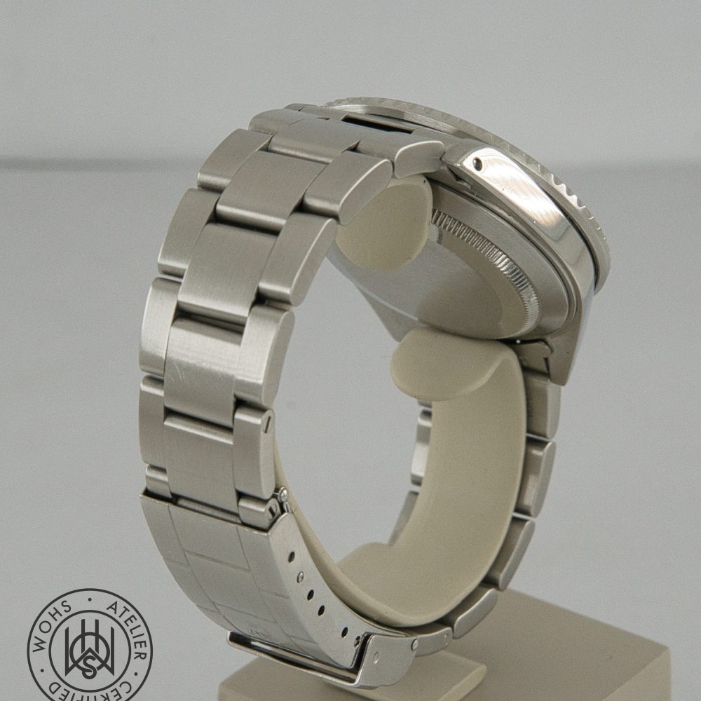 Close-up of a silver stainless steel watch bracelet on a display stand, showing the case and clasp from the side, with a circular certification seal in the bottom left. Rolex Submariner reference 16610 preowned 1998 box papers 4